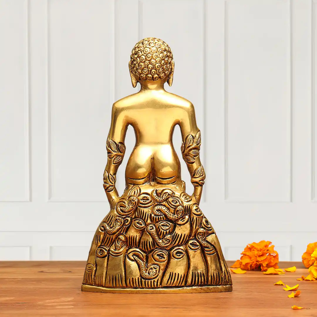 A gold-colored metallic Buddha statue shown from the back, seated with ornate carved details on the draped lower body, placed on a wooden surface against a white paneled wall with orange flower petals nearby.