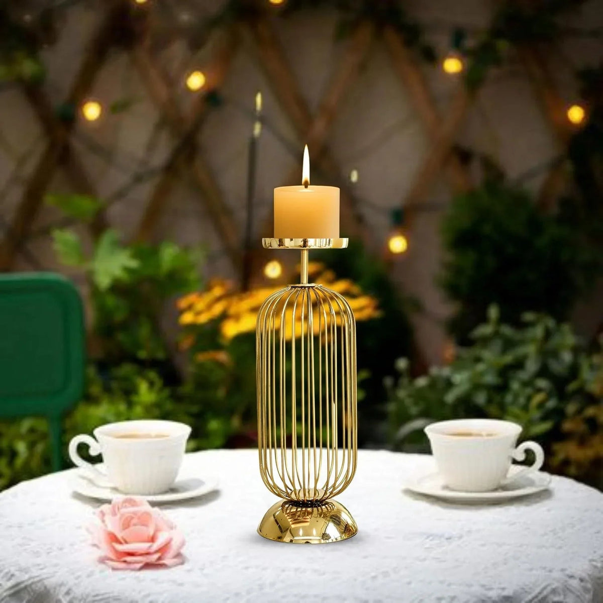 Gold candle holder with a lit candle on a table with two cups and a pink flower, outdoor setting