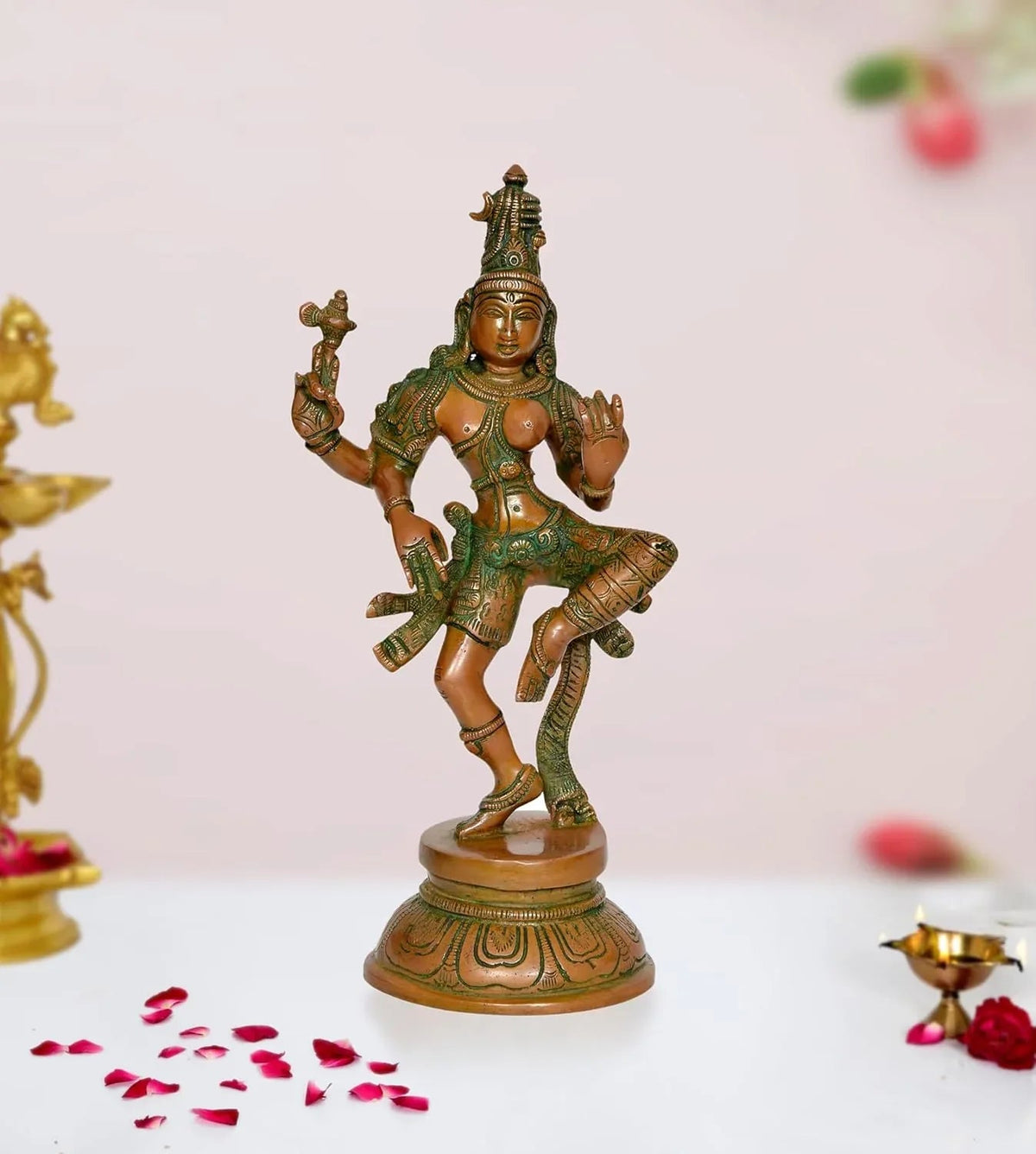 A brass statue of Ardhanarishvara, representing the divine union of Shiva and Parvati, in a South Indian style. The statue is on a base with a circular frame, and there are decorative elements around it, like a crescent moon on Shiva's head and floral details.