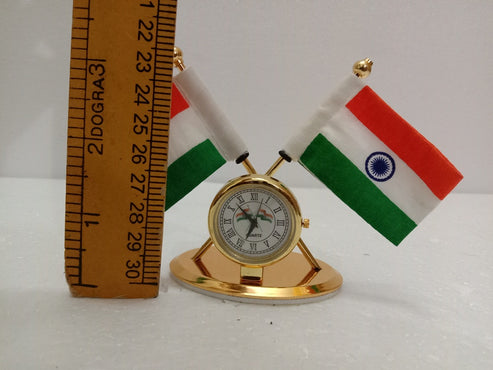 A miniature gold desk clock with two small Indian flags on crossed poles, shown next to a wooden ruler for scale on a white background.