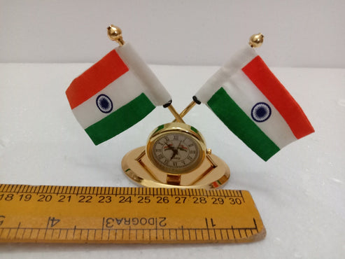 A miniature gold-colored desk clock with two small Indian flags on crossed poles, displayed above a wooden ruler to show scale on a white background.