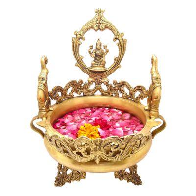 A brass urli (bowl) with a traditional elephant design, featuring a statue of Lakshmi at the top, surrounded by pink rose petals and decorative elements.