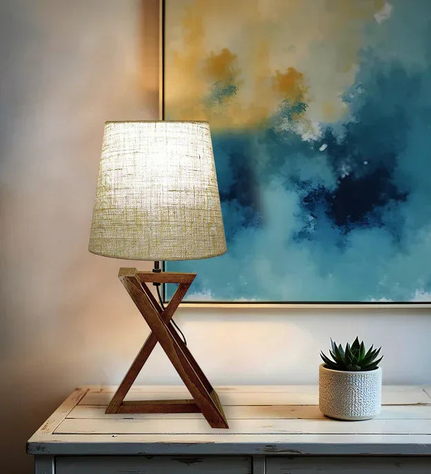 Table lamp with a textured shade on a wooden table, next to a potted plant and a piece of abstract art.