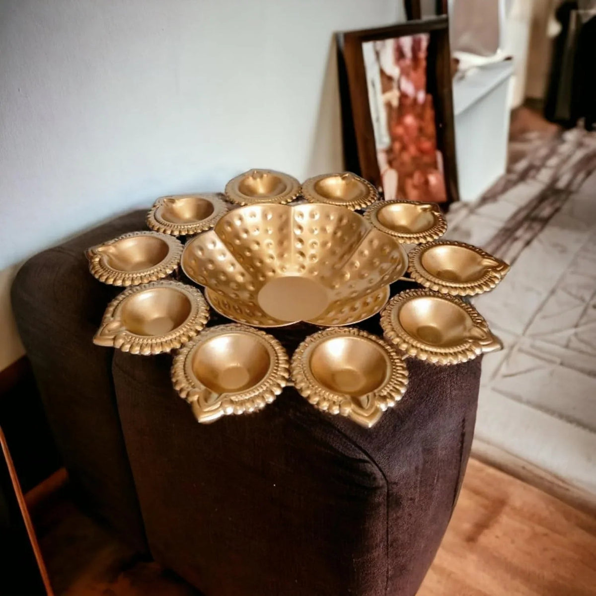 A golden Diya Rangoli Urli bowl with intricate lotus flower motifs, displayed on a dark-colored surface, potentially used for holding diya lamps or as a decorative piece during festivals.