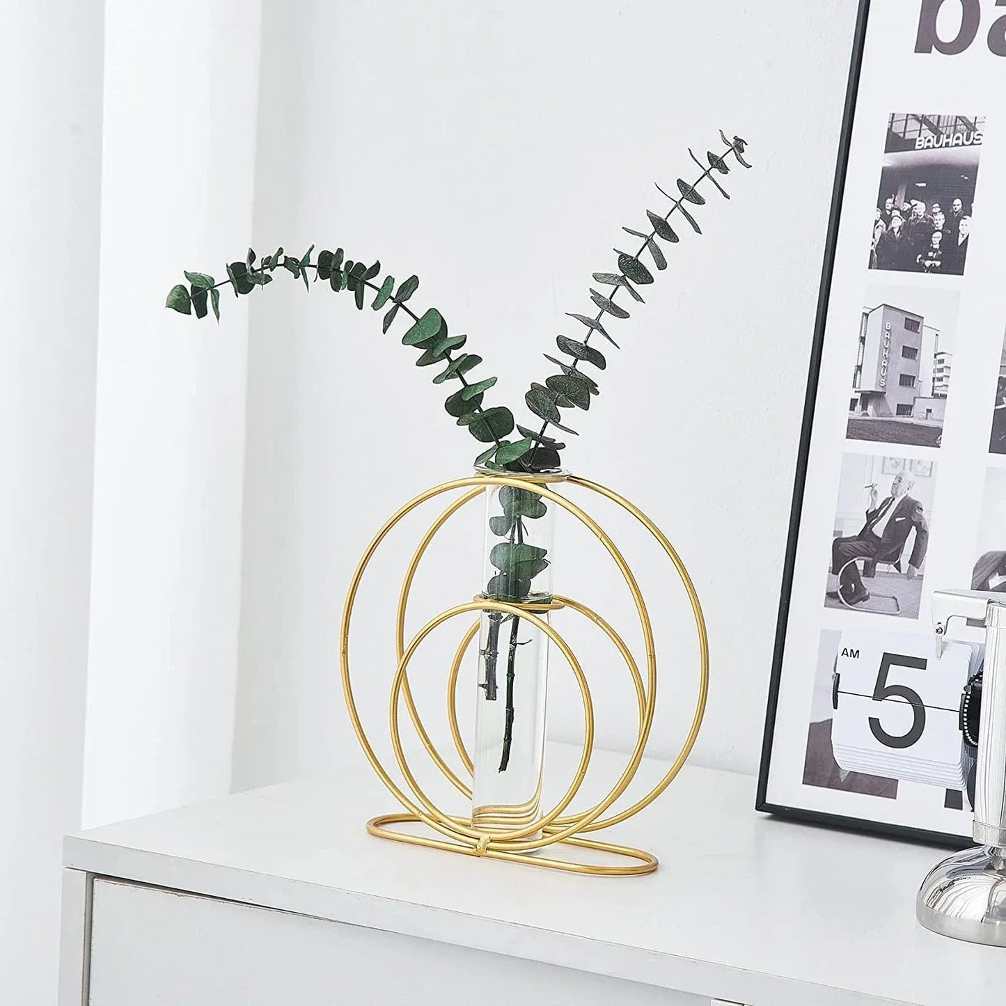 Gold circular vase with a plant on a white surface next to a framed picture