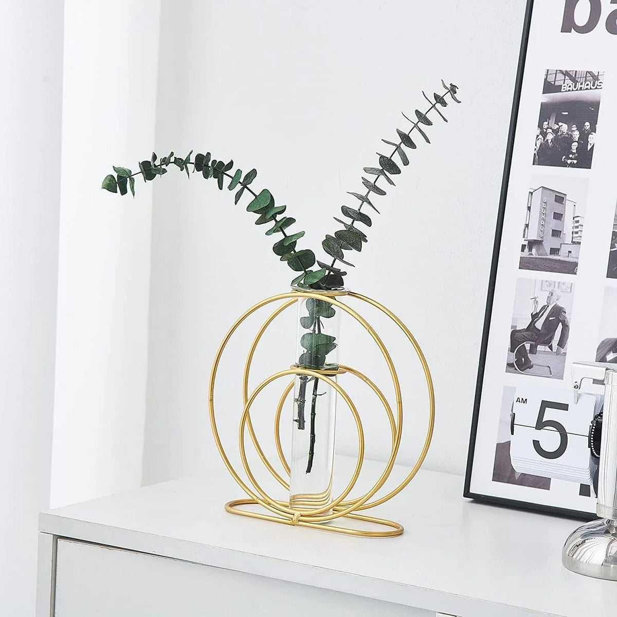 Gold circular vase with a plant on a white surface next to a framed picture