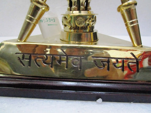 A brass religious trophy or plaque with the engraved Hindi text 'Satyamev Jyate' on the front, featuring two upward-pointing brass prongs and a central decorative finial, mounted on a dark wooden base.