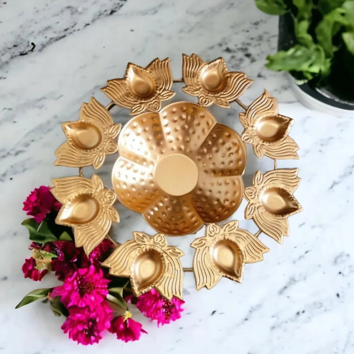A golden circular Lotus Diya Rangoli Bowl with intricate lotus flower designs, placed on a marble surface with pink flowers and a diya lamp in the background.