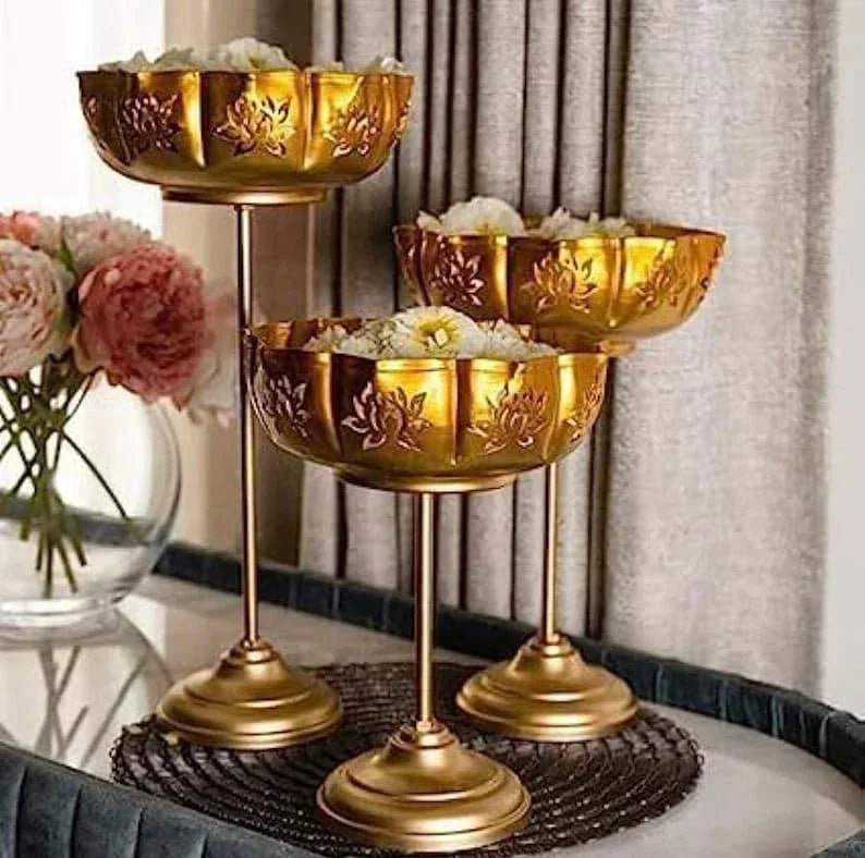 A set of three gold-colored iron urli stands with lotus petal designs, displayed on a textured surface with a floral arrangement in the background.