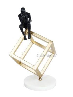 A bronze sculpture of a man sitting in a thinking position on top of a gold-colored geometric frame, with a white base.
