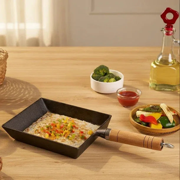 Small black frying pan with food on a wooden table, surrounded by bowls of vegetables and a bottle of oil.