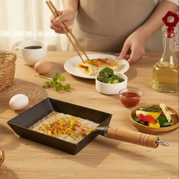 Person preparing food on a wooden table with a frying pan, vegetables, and a bottle of oil.
