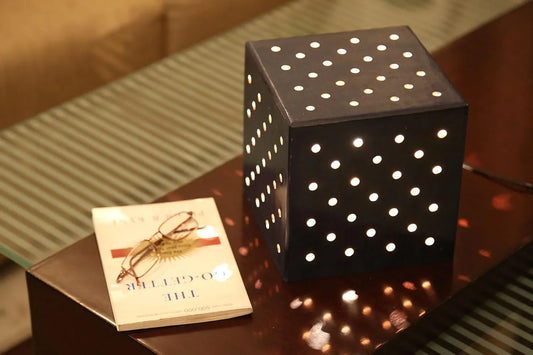 A wooden dice table lamp with a pattern of holes on its base, emitting a warm glow, placed on a surface next to a book.