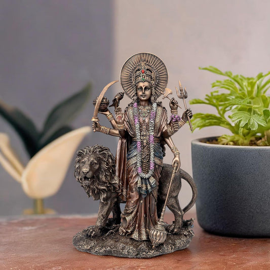A detailed polyresin-finished statue of Goddess Lakshmi standing beside a lion, displayed on a wooden surface with a blurred potted plant in the background.