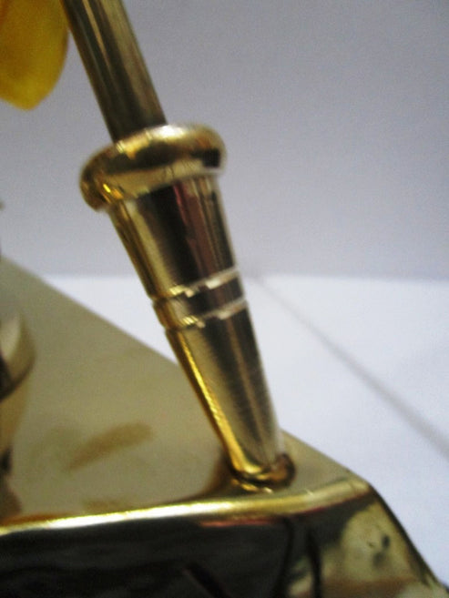 Close-up detail of a polished gold-toned metal object, showing a cylindrical rod mounted on a reflective base against a plain light background.