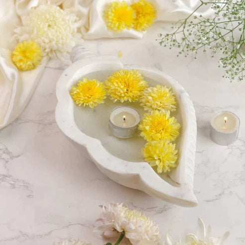 Decorative arrangement with yellow flowers and candles in a heart-shaped white dish on a marble surface.
