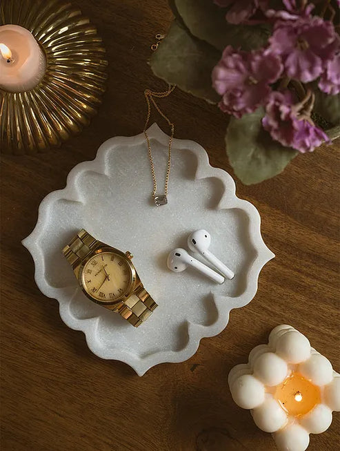 White decorative dish with a gold watch, white AirPods, and a necklace on a wooden surface with candles and flowers.