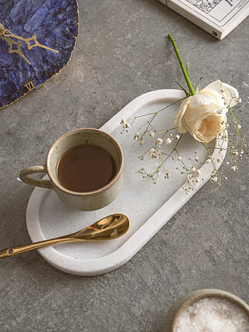 Ceramic mug with coffee, gold spoon, white rose, and marble tray on a gray surface.