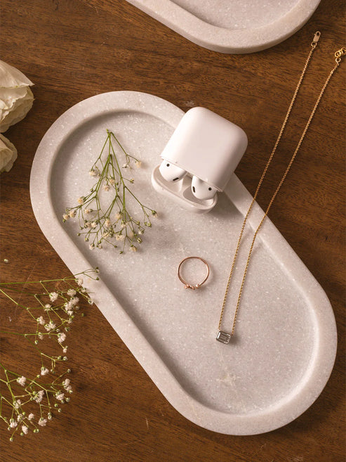 White wireless earbuds on a stone tray with a gold necklace and ring, on a wooden surface.