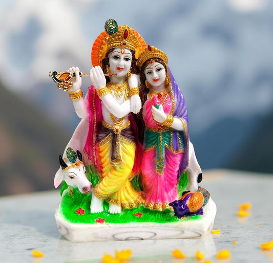 A colorful figurine of Lord Krishna and Rukmi standing on a white marble-like base with a cow at their feet, set against a blurred mountain background.