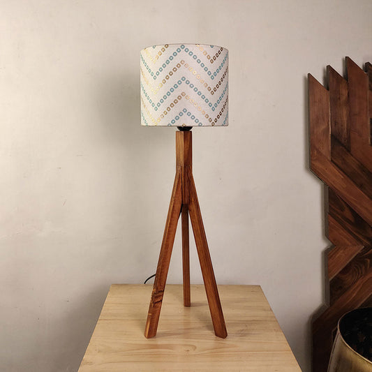 Wooden tripod lamp with patterned fabric shade on a wooden surface against a beige wall.