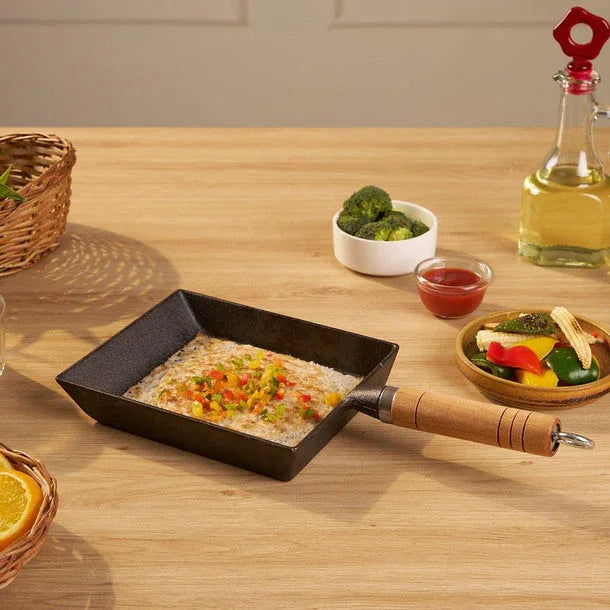 Small black frying pan with food on a wooden table, surrounded by ingredients and a bottle of oil.