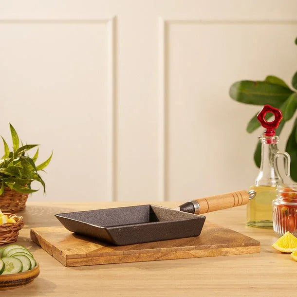 Small black frying pan with wooden handle on a wooden cutting board, with a plant and bottles